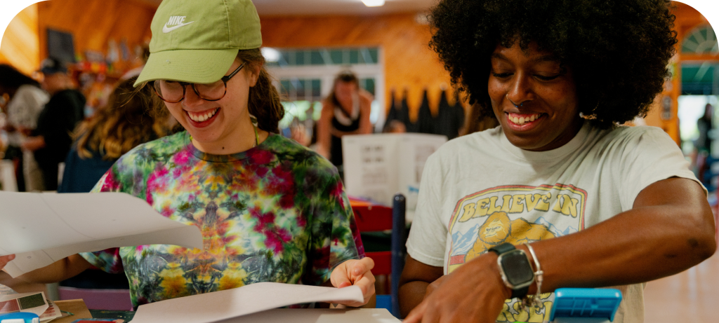 Two women smiling and engaging in a creative activity, surrounded by a lively indoor setting.