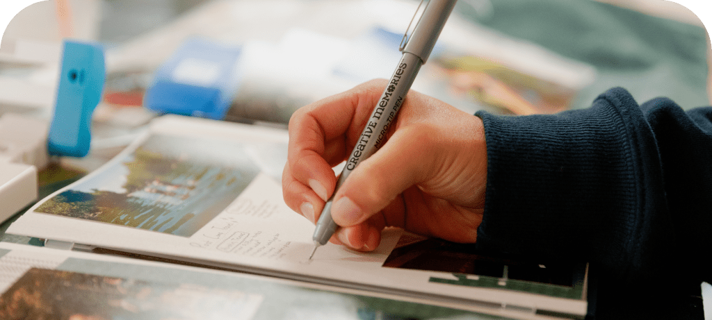 A close-up of a hand holding a gray pen writing notes on a piece of paper, surrounded by photographs and craft supplies.