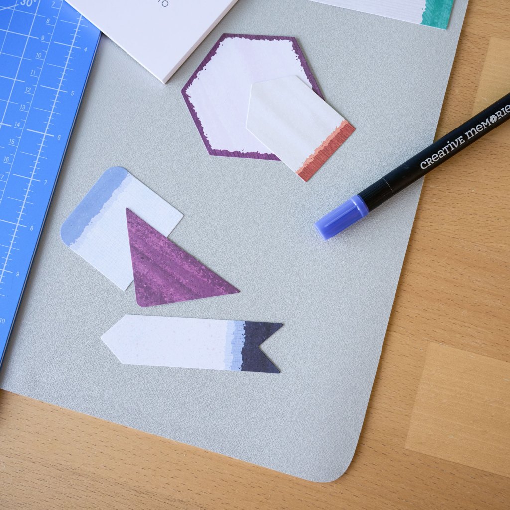 A flat lay of colorful cut-out paper shapes in various designs, including hexagons and banners, on a gray cutting mat. A blue ruler and a purple marker are also visible.