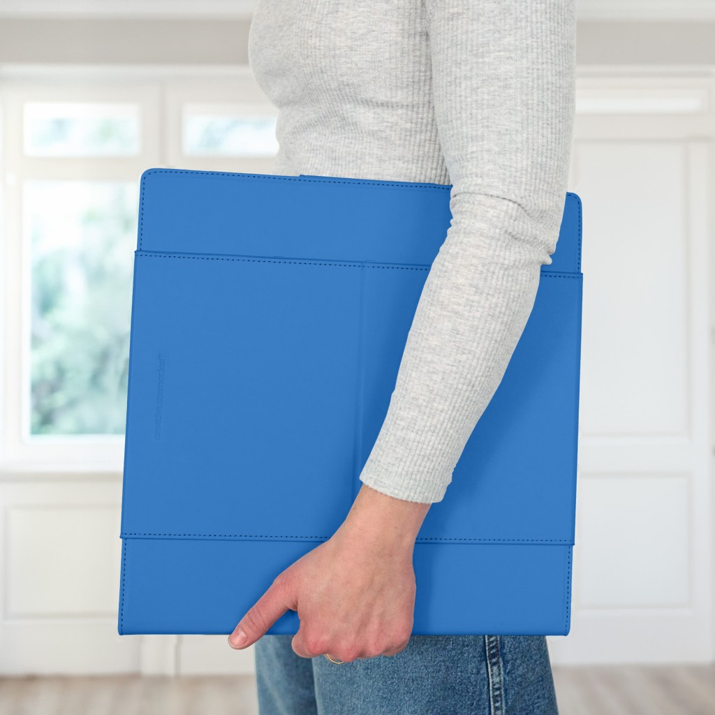 A person holding a bright blue folder or binder, dressed in a gray long-sleeve shirt and jeans, standing in a well-lit room with large windows.