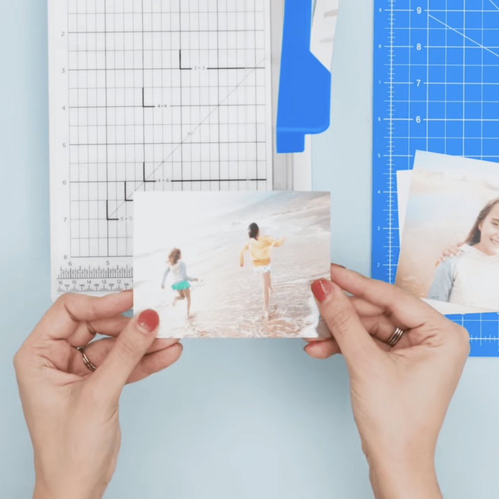 A person holding a photograph of two children running on the beach, with crafting tools and a cutting mat in the background.
